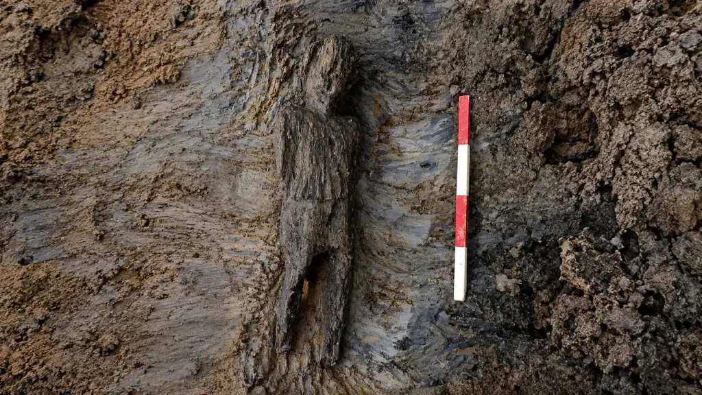 Obra de ferrovia abre vala encharcada em um campo e encontra figura humana de carvalho com 67 centímetros, túnica e músculos esculpidos, preservada por quase 2 mil anos e cercada por mistério sobre possível ritual romano