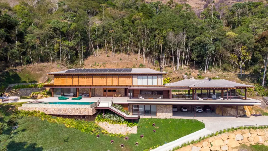 Casa de 1.200 m² nasce num terreno íngreme de 10 mil m² em Itaipava e, durante a obra, arquitetos encontram uma rocha gigante, decidem não removê-la, moldam vidros à geometria da pedra e incorporam o achado à sauna do spa, enquanto outras rochas passam a apoiar a garagem e a construção