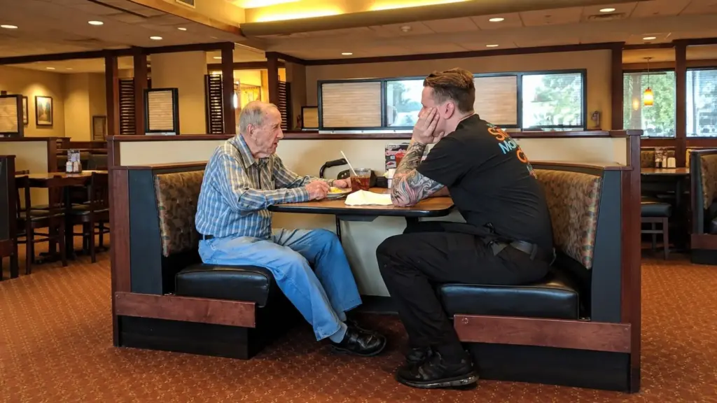 Garçom atende veterano de 91 anos que havia esquecido os aparelhos auditivos, ajoelha-se para facilitar a conversa e escuta o cliente revelar que vivia sozinho e quase não tinha com quem falar, então usa o próprio intervalo para sentar à mesa e acompanhar o idoso durante toda a refeição