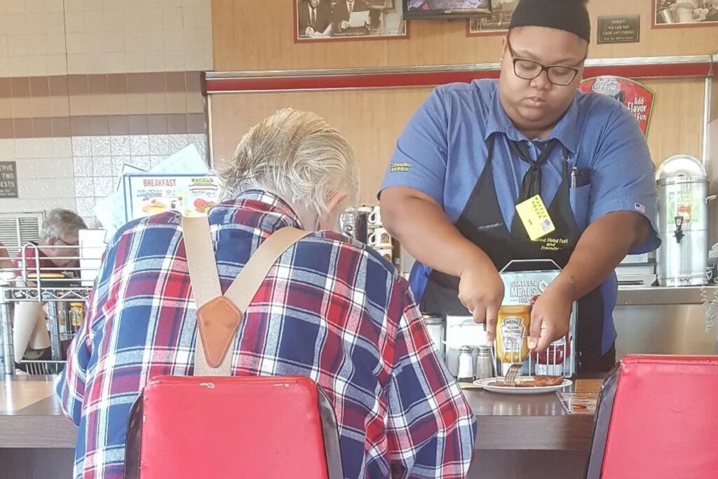 Garçonete de 18 anos percebe que cliente idoso recém-operado e com tanque de oxigênio não conseguia cortar a própria comida, para o atendimento para cortar o presunto dele pedaço por pedaço e se emociona quando a foto do gesto rende a ela uma bolsa de estudos de cerca de R$ 83 mil