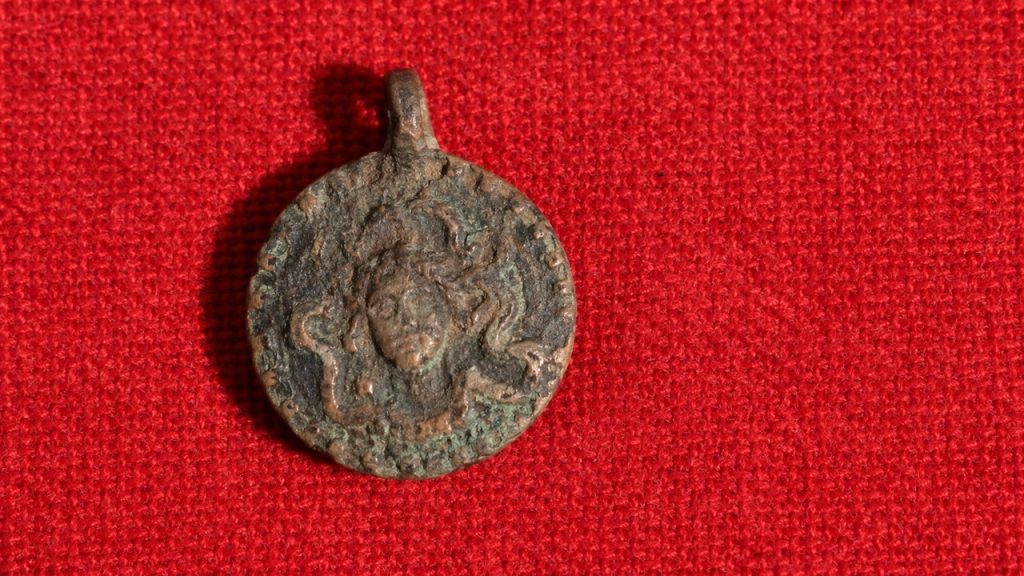 Arqueólogos encontram peça de cobre com santo guerreiro de um lado e figura semelhante à Medusa cercada por serpentes do outro, em escavação medieval, revelando como a fé cristã e crenças pagãs coexistiam há 1.000 anos